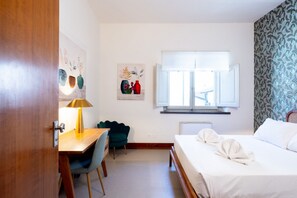 Habitación de hotel con cama, escritorio con lámpara, silla, ventana con persianas y una obra de arte en la pared.