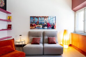 Un salon moderne avec un grand tableau, deux canapés gris ornés de coussins rouges, une petite table d’appoint et un lampadaire.