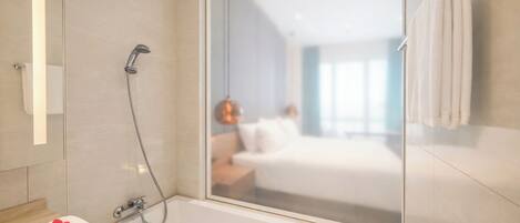 A modern bathroom with a glass shower enclosure, a bathtub, and a visible bedroom with a bed and nightstand.
