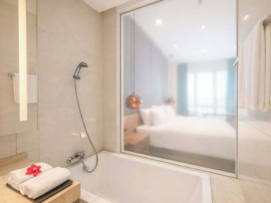 A modern bathroom with a glass shower enclosure, a bathtub, and a visible bedroom with a bed and nightstand.