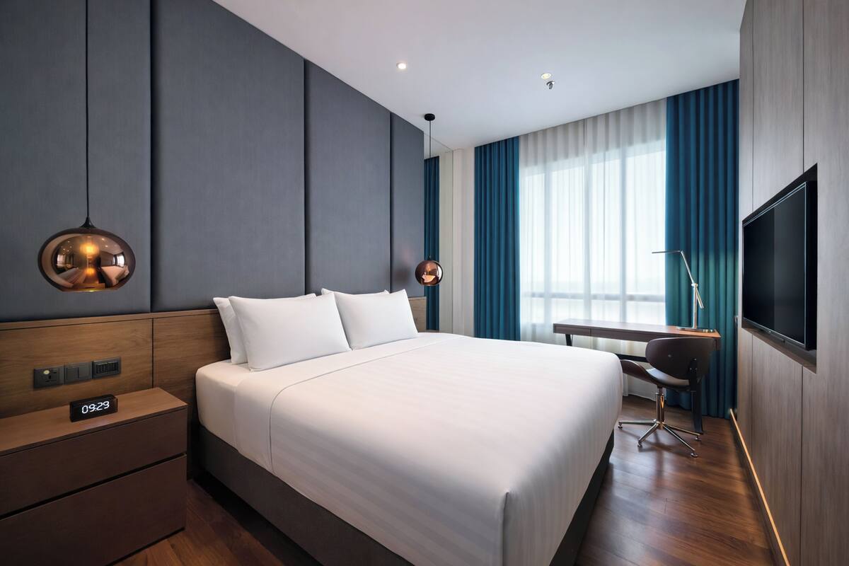 Ein modernes Hotelzimmer mit einem großen Bett, Bedienelementen am Bett, einem Schreibtisch mit Stuhl, einem Fernseher und einem Fenster mit Vorhängen.