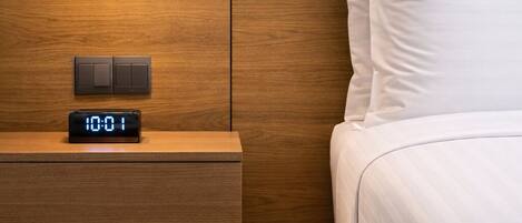 A modern hotel room with a wooden nightstand, a digital clock, a bed with white bedding, and a unique spherical pendant light.