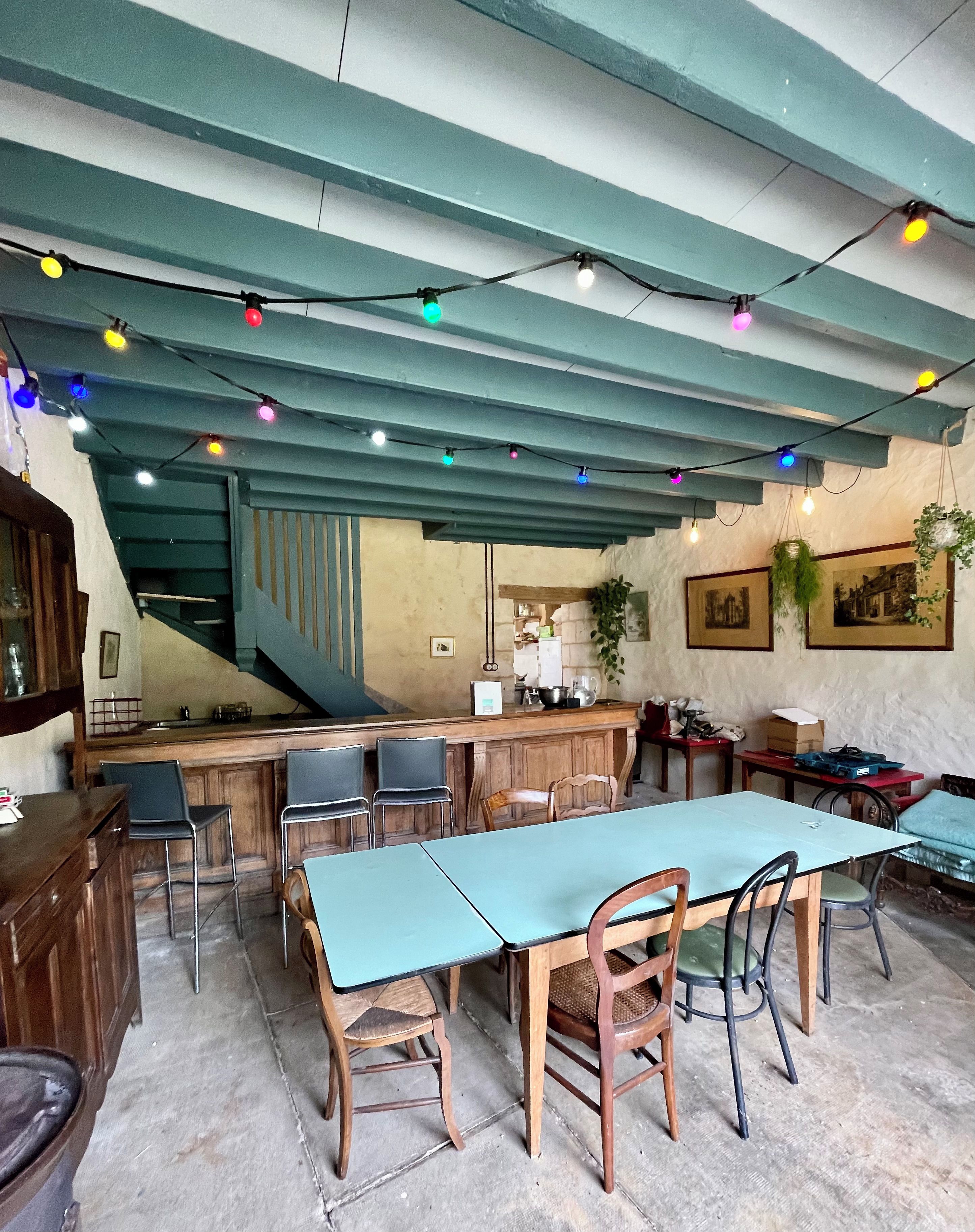 Un espace repas avec une longue table, des chaises et un comptoir de bar. Des guirlandes lumineuses sont suspendues au plafond et un escalier se trouve au fond de la pièce.
