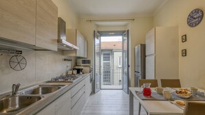 Una cuina moderna amb electrodomèstics d'acer inoxidable, una zona de menjador amb taula preparada per a dos i una vista a un balcó per una porta oberta.