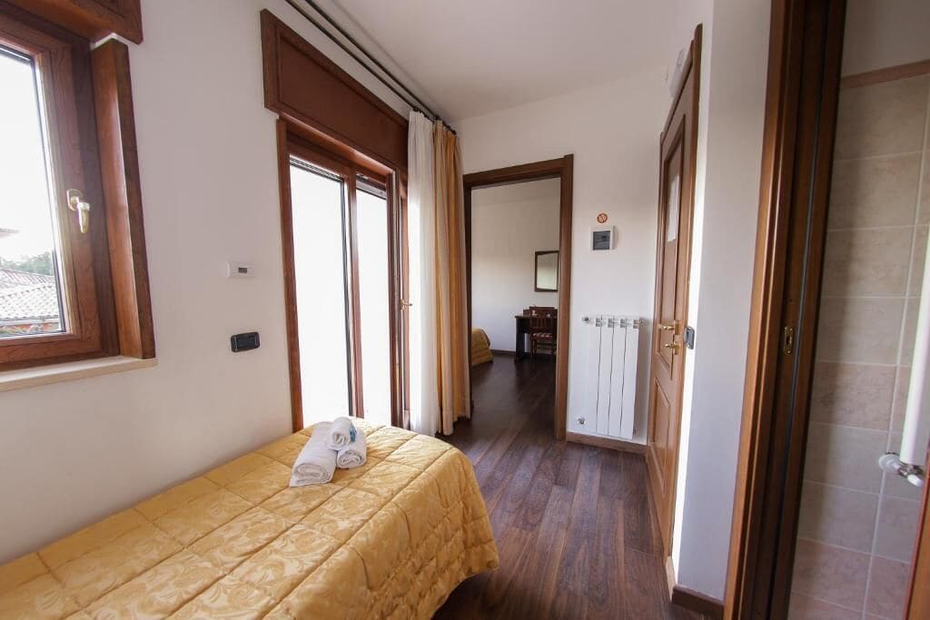 Ein Hotelzimmer mit Bett, Fenster, Tür und einem Flur, der zu einem weiteren Zimmer führt.