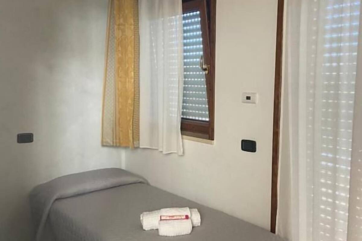 A single bed with a white bedspread, a window with blinds, and a curtain rod with a yellow curtain.