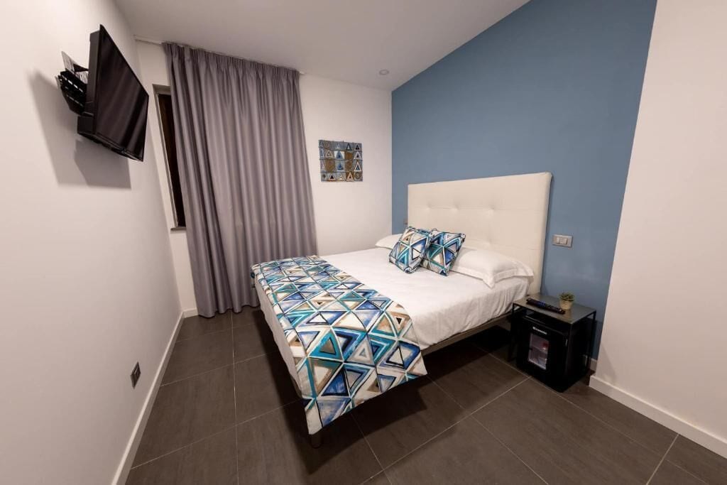 Een slaapkamer met een bed, gordijnen, een aan de muur bevestigde televisie, een nachtkastje en een kleine plant.
