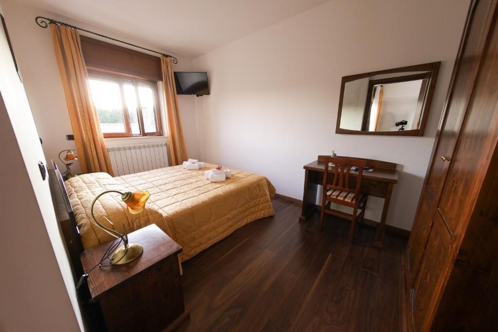 Un dormitorio con cama, mesita de noche, silla, escritorio, espejo, televisor y ventana con cortinas.