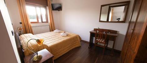 Un dormitorio con cama, mesita de noche, silla, escritorio, espejo, televisor y ventana con cortinas.