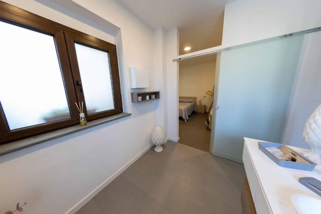 A hotel room with a large window, a small decorative item on the floor, a white bedside table, and a view into the adjacent room.