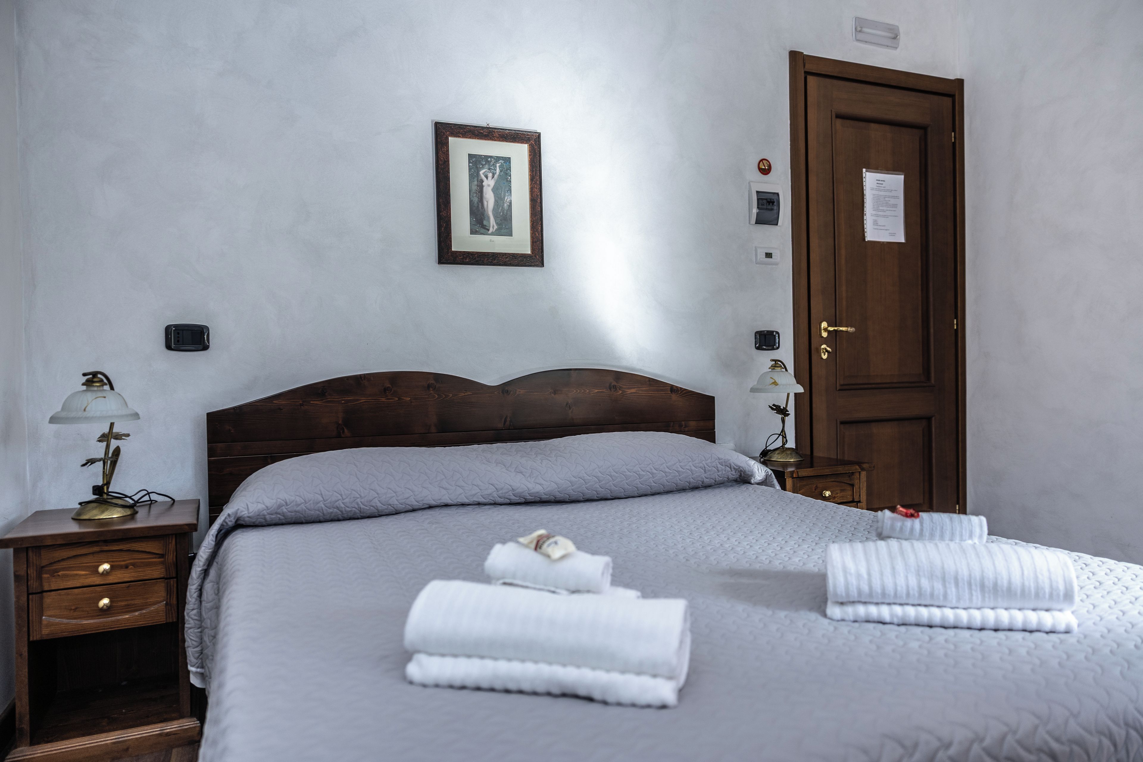 Ein Hotelzimmer mit einem hölzernen Kopfende, einem Bett mit grauer Decke, zwei Nachttischlampen, einem Nachttisch, einer Tür mit einem Schild und einem gerahmten Bild an der Wand.