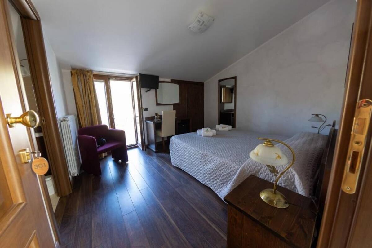 A hotel room with a bed, a desk with a lamp, a chair, a window, and a door with a number.