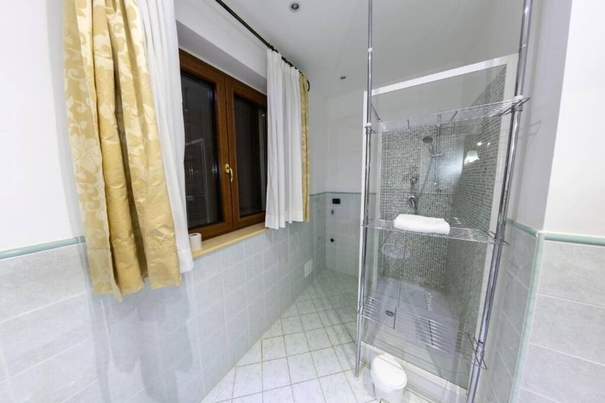 A bathroom with a glass shower enclosure, a toilet, a window with curtains, and tiled walls.