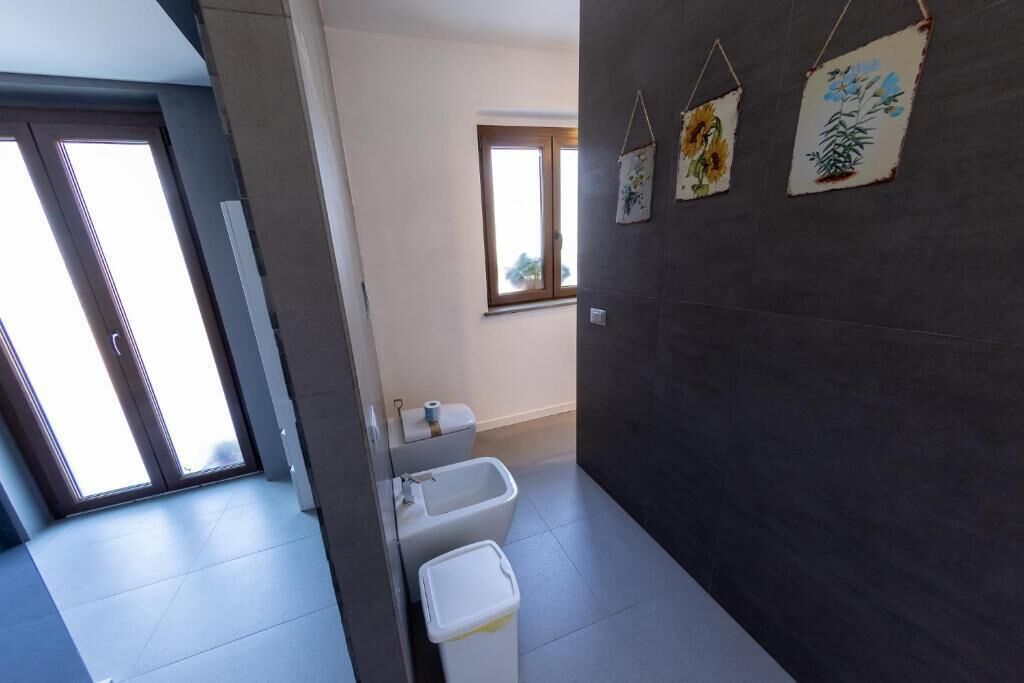 A bathroom with a toilet, bidet, and trash bin, a window with a view, and a door leading to another room.