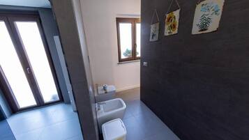 A bathroom with a toilet, bidet, and trash bin, a window with a view, and a door leading to another room.