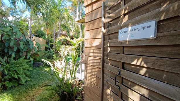 Una verja de madera con un cartel que dice 'Cabane des tropiques' y abundante vegetación tropical.