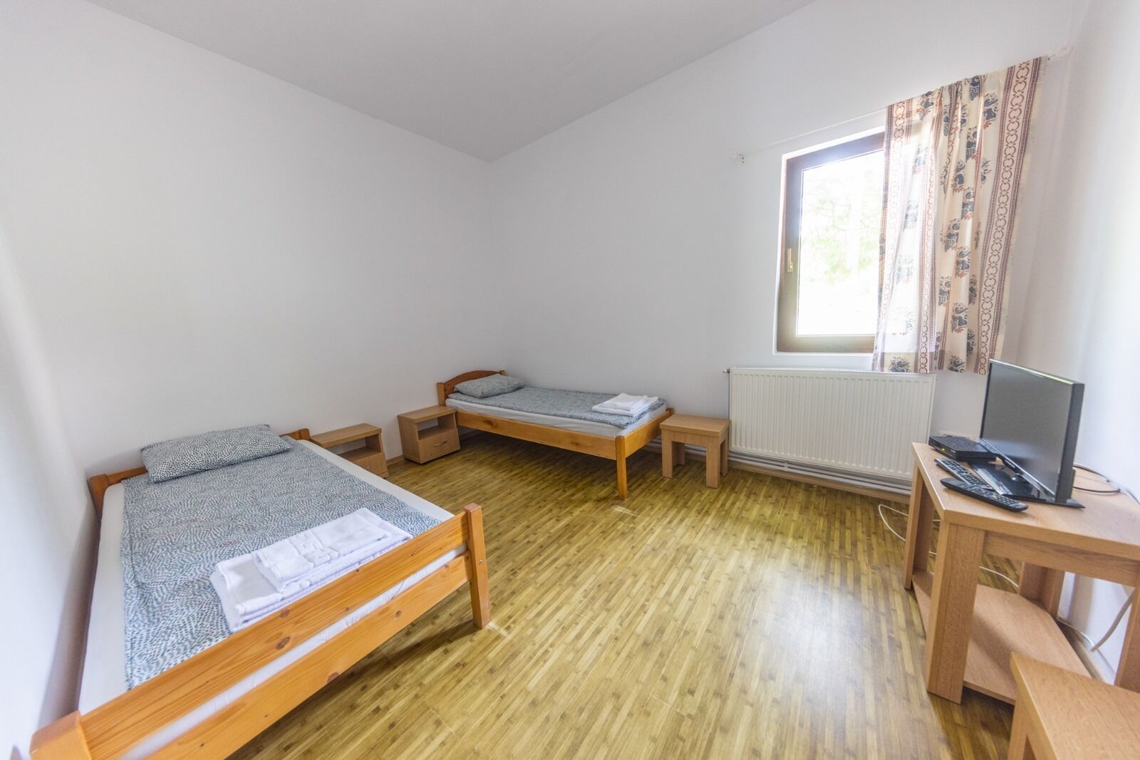 A small, single-room accommodation with two wooden beds, a desk with a computer, and a window with curtains.