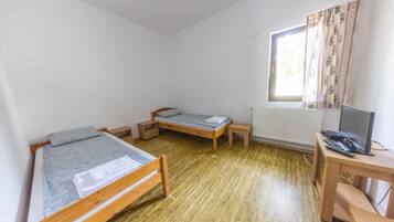 A small, single-room accommodation with two wooden beds, a desk with a computer, and a window with curtains.
