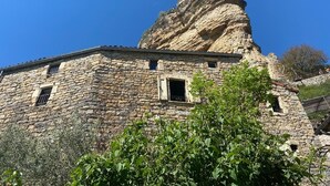 A stone building with a prominent rock formation on top, a window, and greenery in the foreground.