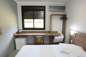 Une chambre d’hôtel comprenant un lit, un bureau avec une chaise, un petit réfrigérateur, une fenêtre avec des stores et un porte-manteau.