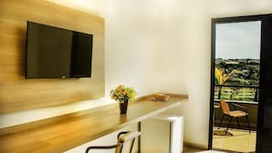 A modern living room with a flat-screen TV mounted on a wooden wall, a small table with a potted plant, a chair, and a sliding glass door leading to a balcony with a view of a green landscape. - HOtel Recreio São Jorge (Jaboticabal)