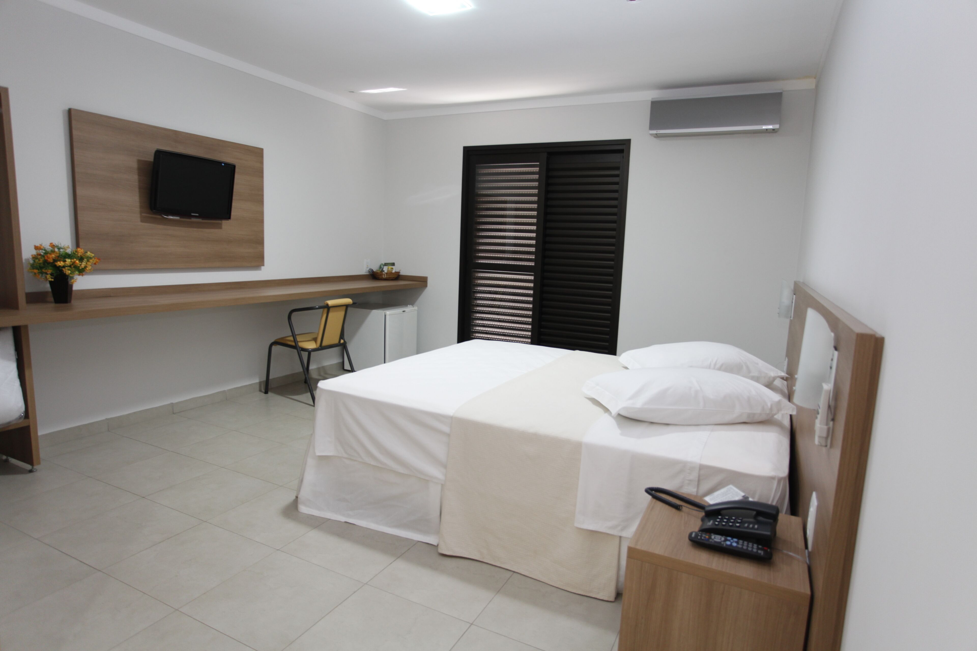 A hotel room with a bed, a desk with a chair, a TV mounted on the wall, and a window with blinds.