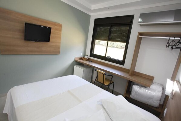 A hotel room with a bed, a desk, a chair, a TV mounted on the wall, a window with blinds, and a wardrobe.