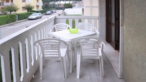 A balcony with a white table and chairs, a green basket, and a view of a street with parked cars and buildings. - Reliable Family Apartment in a Great Area (Lignano Sabbiadoro)