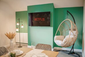 A dining area with a table set for two, a hanging chair, a floor lamp, and a wall-mounted television displaying 'NETFLIX'. - Velvet Residence Central Sheffield | Parking 12PPL (Sheffield)