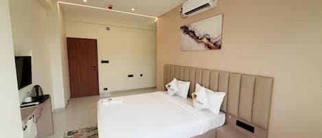 A modern hotel room with a large bed, a flat-screen TV, a desk with a kettle, and an air conditioning unit.