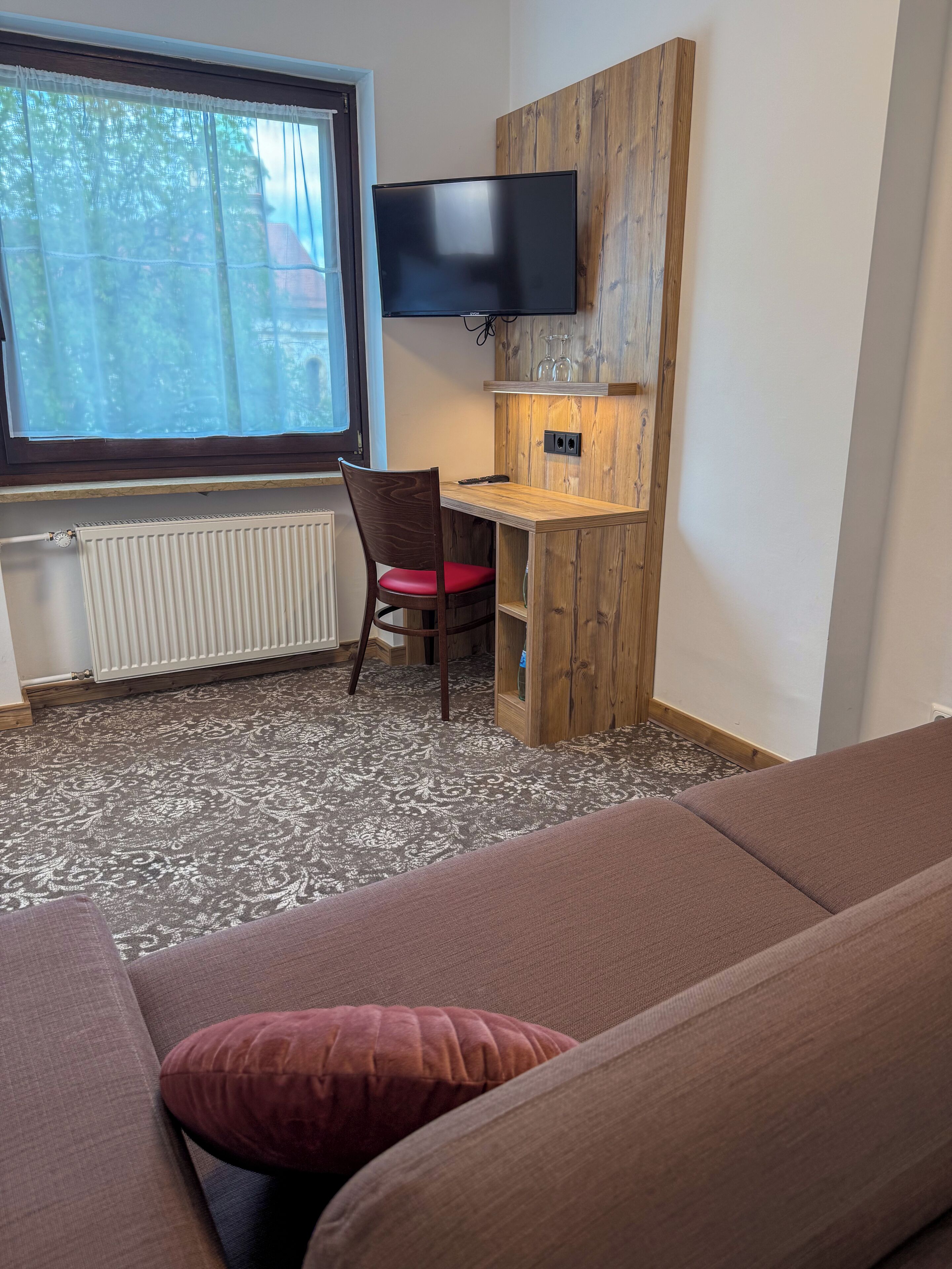 A room with a sofa, a wooden desk, a chair, a television, and a window with a view of trees.