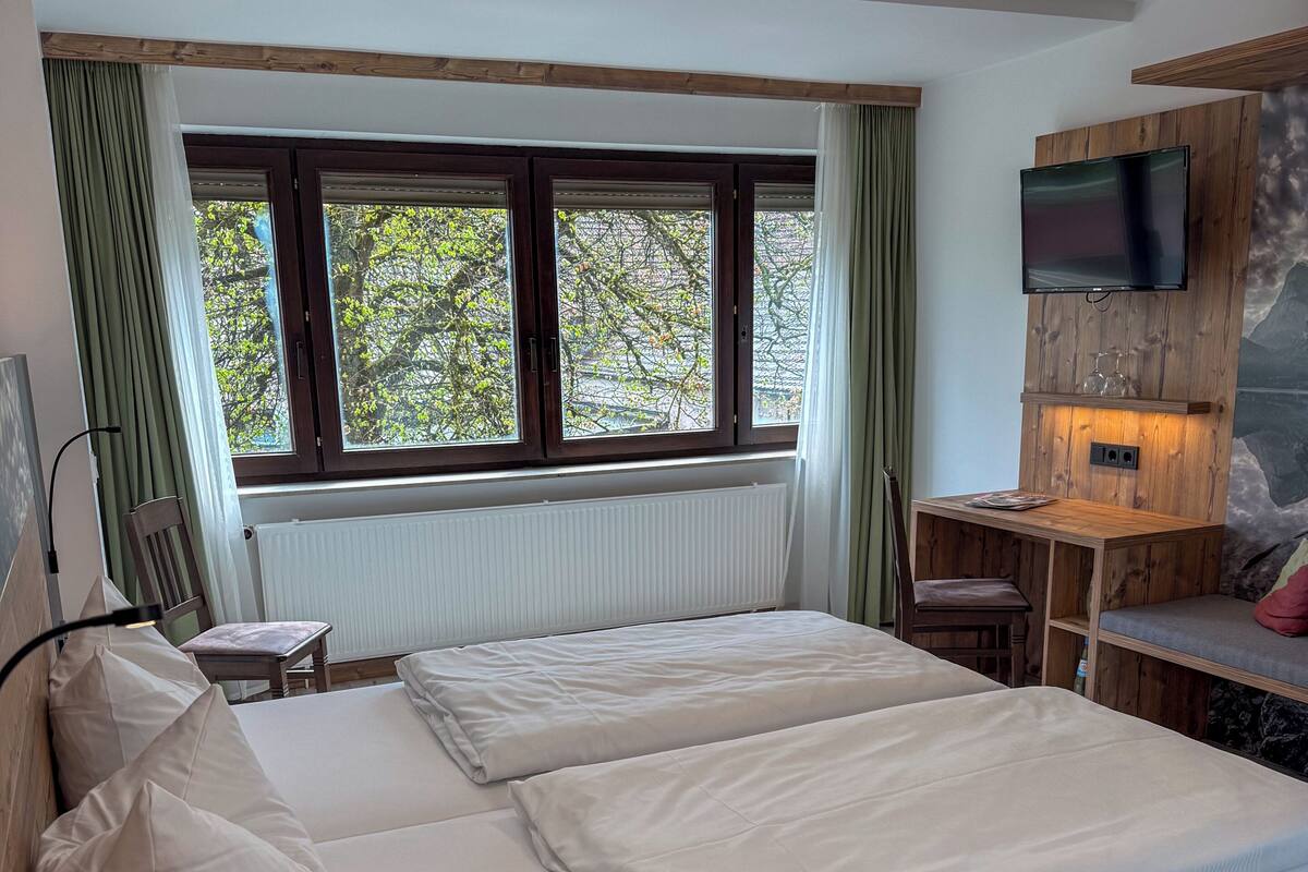 Ein Schlafzimmer mit Bett, Schreibtisch, Stuhl, Fernseher und einem Fenster mit Vorhängen.