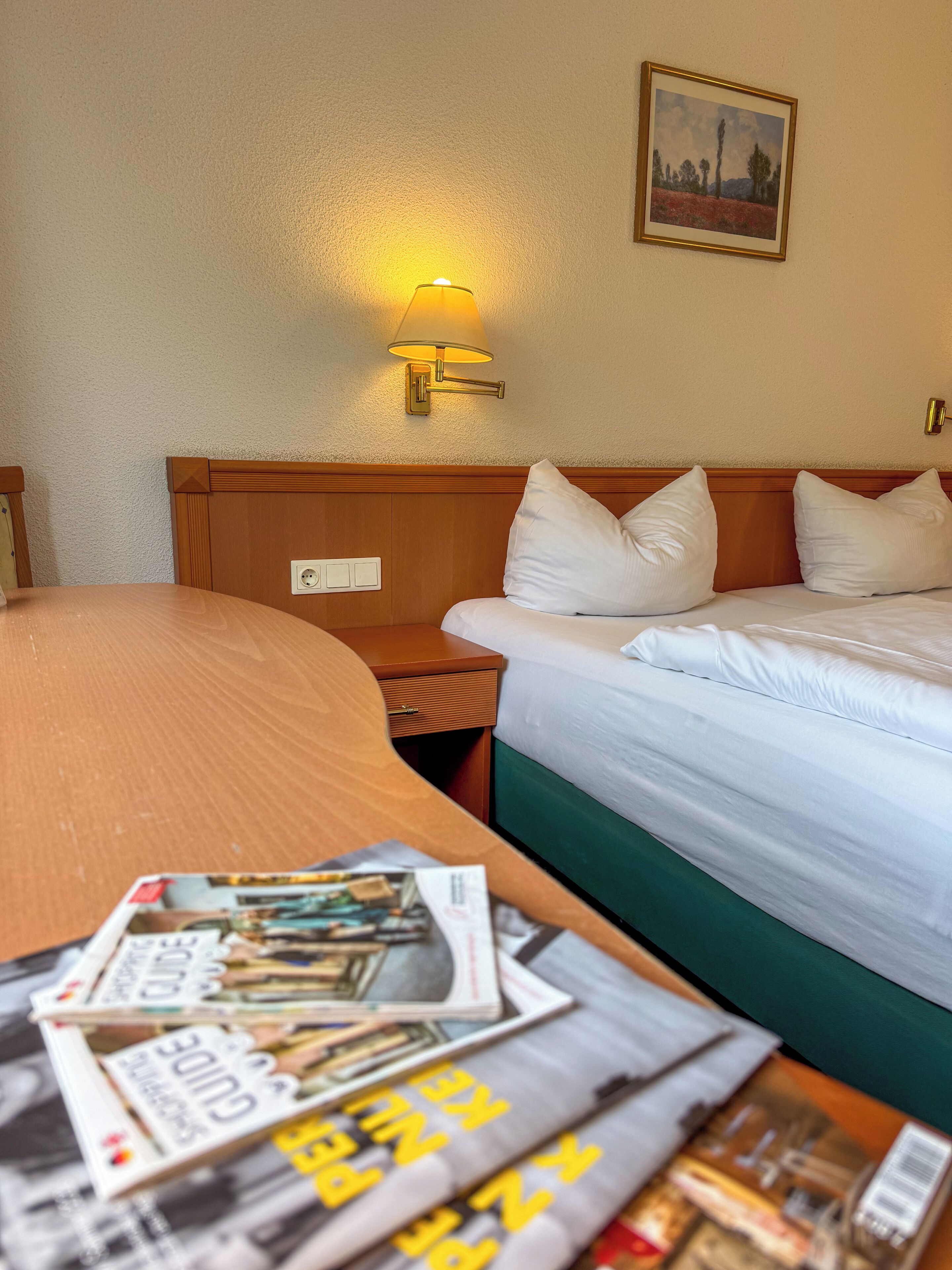 A hotel room with a bed, bedside table, lamp, and a wall-mounted painting.