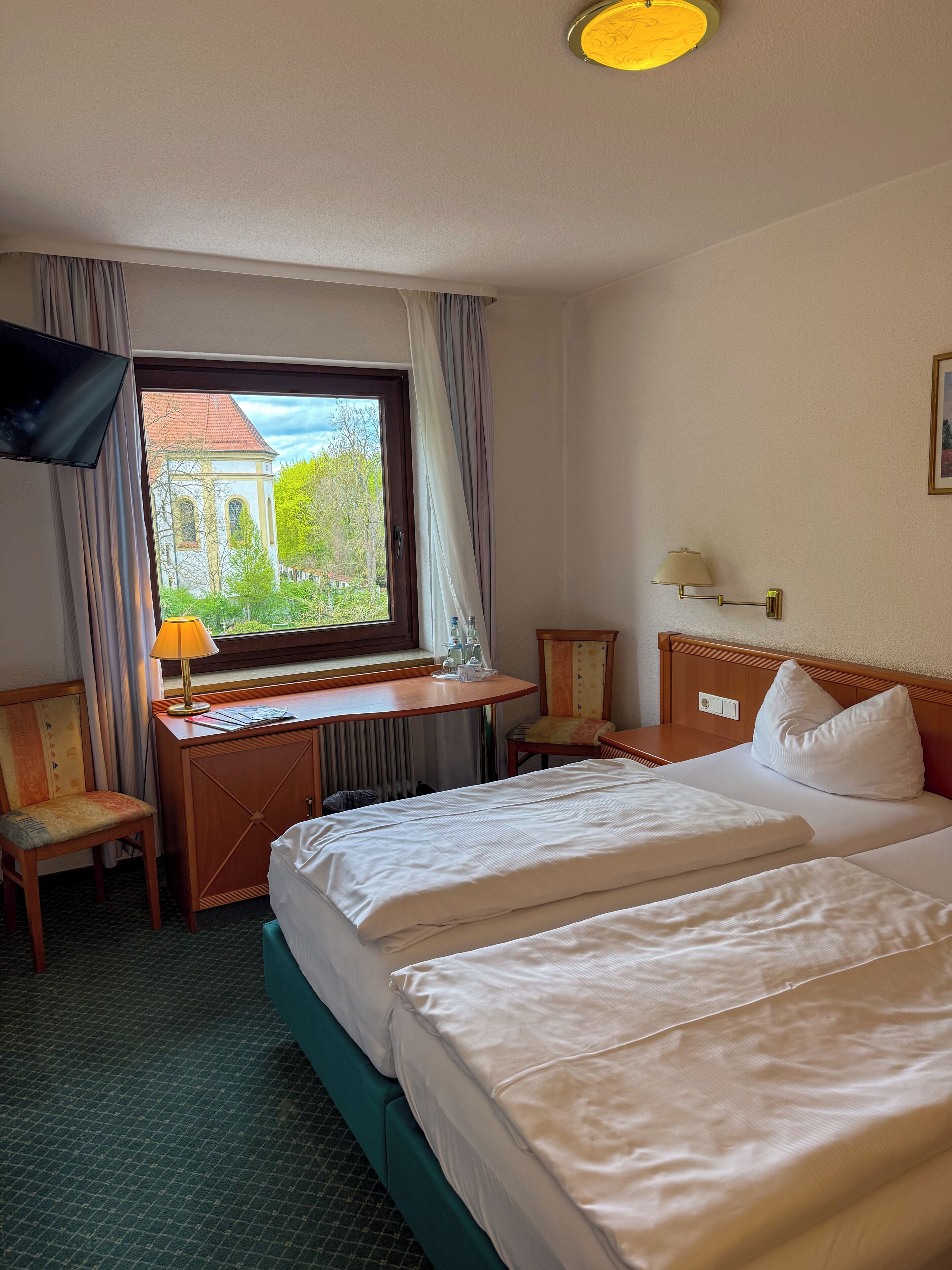 A hotel room with a bed, a desk, a chair, a TV, and a window with a view of a building.