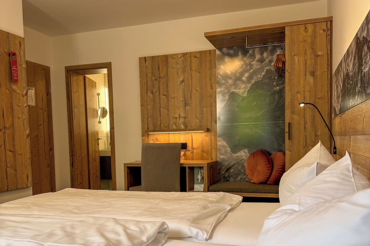 Ein Hotelzimmer mit zwei Betten, Holzwandverkleidung, einer Wandmalerei mit Bergblick und einem kleinen Tisch mit Lampe.