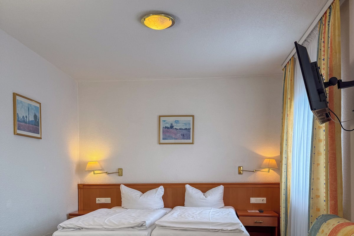 Ein Hotelzimmer mit Doppelbett, Einzelbett, Sofa, zwei Nachttischlampen, einer Deckenleuchte und zwei gerahmten Bildern an den Wänden.