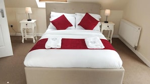 A neatly made bed with a red and white color scheme, two bedside tables with lamps, and a radiator on the wall. - House Sleeps 7 With Garden and Parking (Gorebridge)