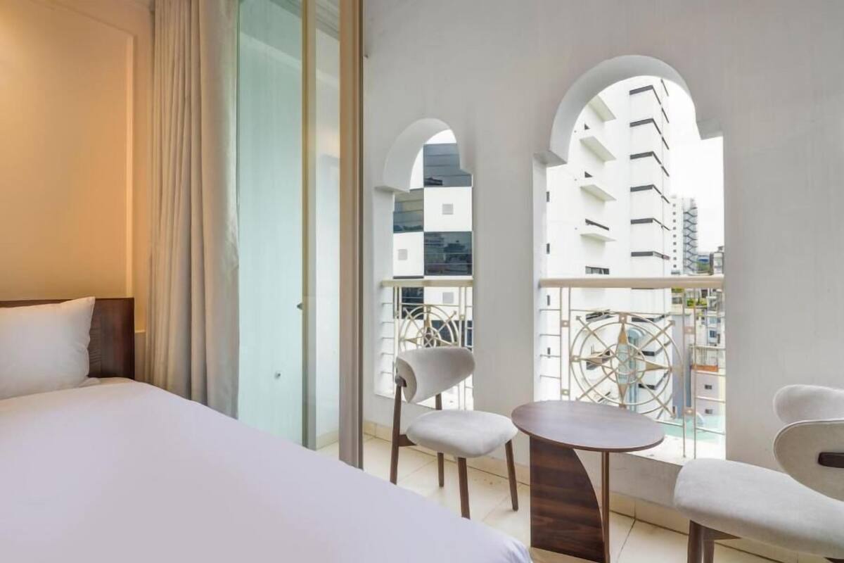 Een hotelkamer met een bed, een tafeltje, twee stoelen en een balkon met uitzicht op gebouwen.
