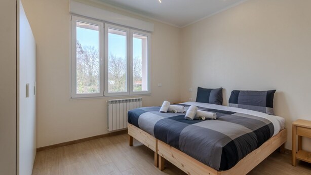 A bedroom with a large bed, a window, a radiator, and a wooden floor. - A bright and welcoming 90 square meters apartment, perfect for seven people, located on the first floor of a building without an elevator.The property is located in Tessera, a suburb close to Marco Polo Airport. The location is also ideal for reaching Ve (Venice)