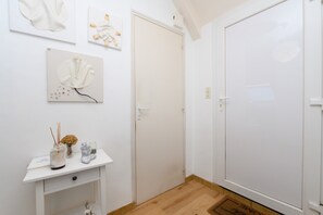 A white room with a wooden floor, a white door, a white wall with three framed artworks, a small white table with a vase and a candle, and a white cabinet with a drawer. - Apartment 'Sarzeau City Center on Foot' with Wi-Fi and Air Conditioning (Sarzeau)