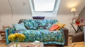 A cozy room with a colorful patterned sofa, a small table with books, a window with a view, and a lamp on a side table. - Apartment 'Sarzeau City Center on Foot' with Wi-Fi and Air Conditioning (Sarzeau)