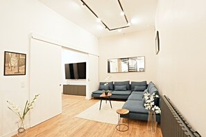 Ein modernes Wohnzimmer mit einer Eckcouch, einem Couchtisch und einem Fernseher. Ein Flur führt zu einer Küchenzeile.