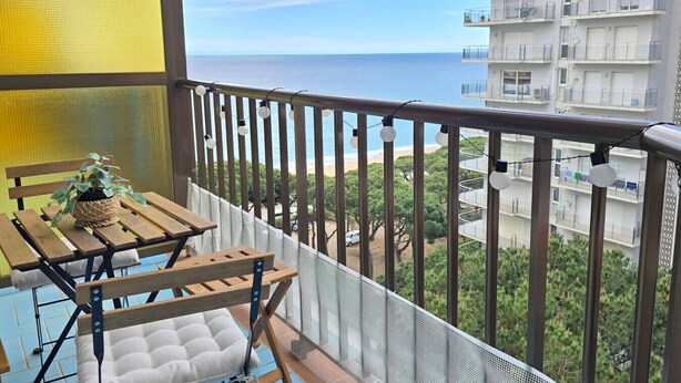 A balcony with a wooden table, a cushioned chair, and a potted plant. - Luminous apartment with two balconies and swimming pool in front of the sea (Blanes)