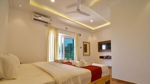 Una habitación de hotel con cama, ventilador de techo, unidad de aire acondicionado, ventana con cortinas y televisor montado.