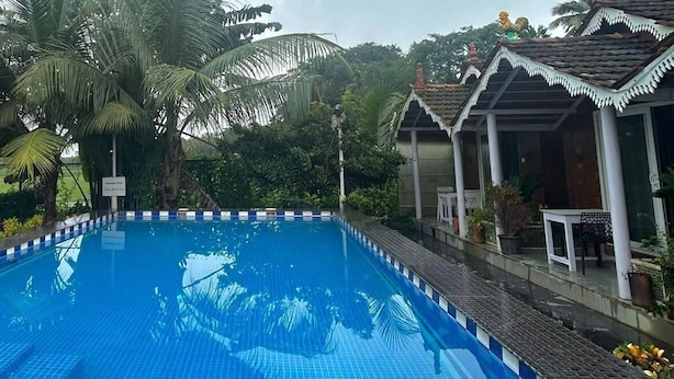 Una piscina con bordes de azulejos, rodeada de abundante vegetación y un edificio con techo de tejas.