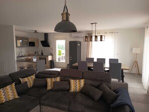 Una sala de estar moderna con un sofá modular, un comedor con mesa y sillas, una cocina con estufa y armarios, y un ventanal grande con cortinas.