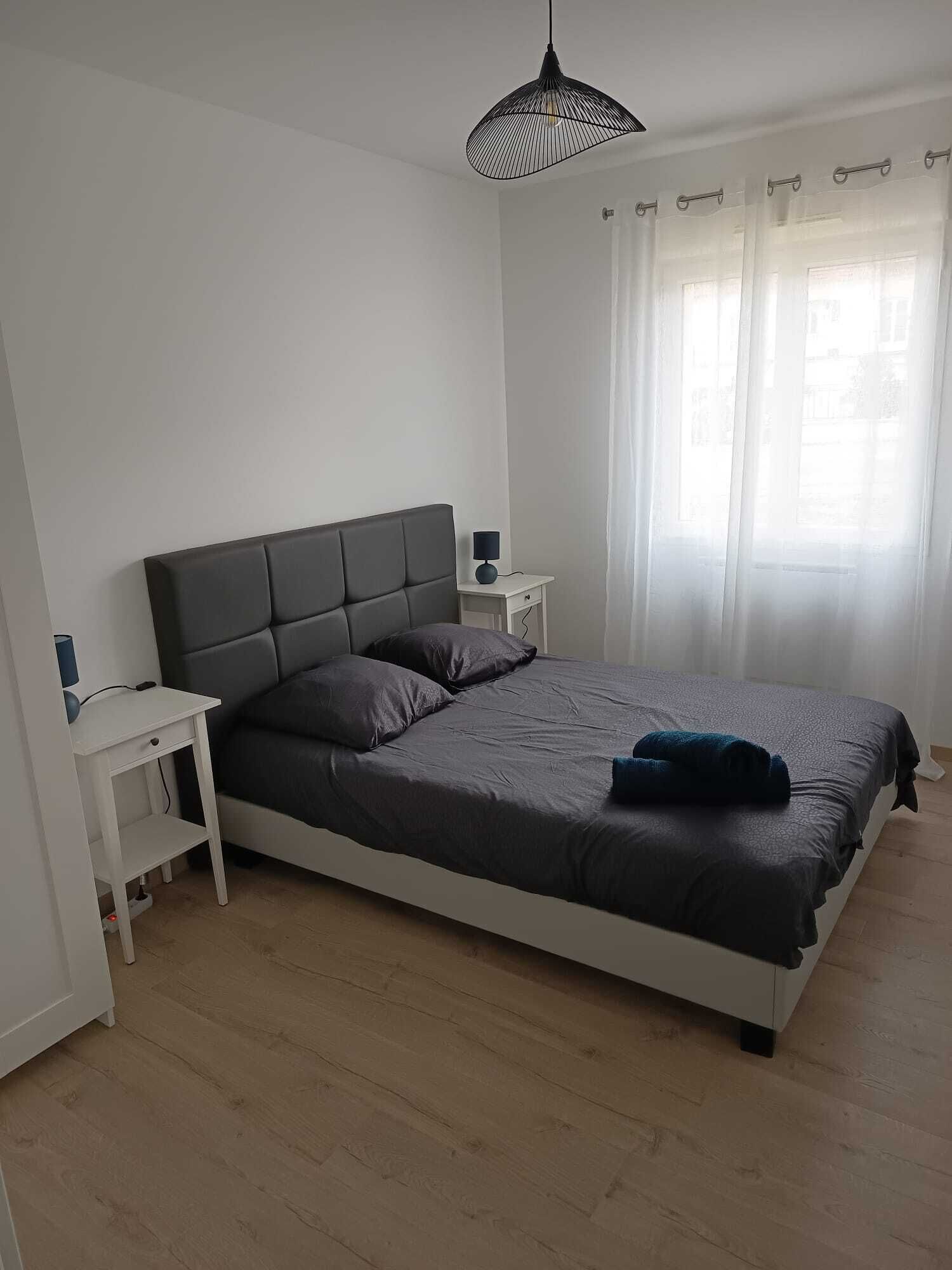 A bedroom with a bed, bedside tables, a lamp, a hanging light fixture, and a window with curtains.