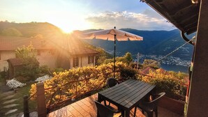 A terrace with a table and chairs, a sun umbrella, and a view of a valley with buildings and mountains. - Little Gem Challet - Trekking House Como Lake (Torno)