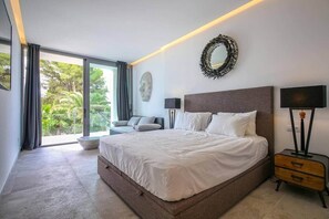 A modern bedroom with a large bed, a sofa, a round mirror, and a nightstand. - Exclusive Villa in the Southwest of Mallorca (Santa Ponsa)