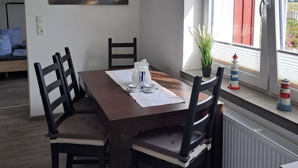 A dining area with a table set for four, chairs, a painting on the wall, and a window with a view. - Seagull's nest in the middle of the countryside (Dornum)
